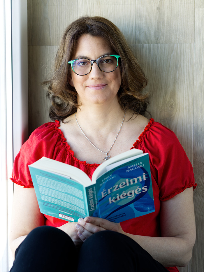 Ez egy fotó: Merényi Katalin Gabriella matematika tanár, life és business coach az érzelmi kiégésről szóló nyitott könyvvel a kezében. Barna, hullámos haja vállig ér, szemüvegkerete zöld-barna, mosolyog. Nyakában egy ezüst láncon ezüst medál kolibri, ami a gondolat kiszabadulásának szimbóluma. A hölgy piros felsőt visel és fekete nadrágot, de csak térdeit látni, mert lábait felhúzva ül egy falnak támaszkodva.