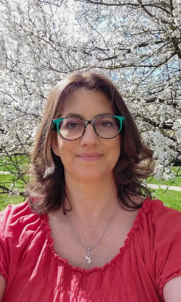 Merényi Katalin Gabriella matematika tanár, life és business coach, art coach mosolyog egy virágzó cseresznyefa előtt. A hölgynek barna, vállig érő hullámos haja van. Szemüvegkerete zöld-barna. Nyakában egy ezüst kolibri medál, mint a gondolat felszabadulásának szimbóluma. Gabriella piros felsőt visel. A fa fehér virágú, zöld ápolt fűben.