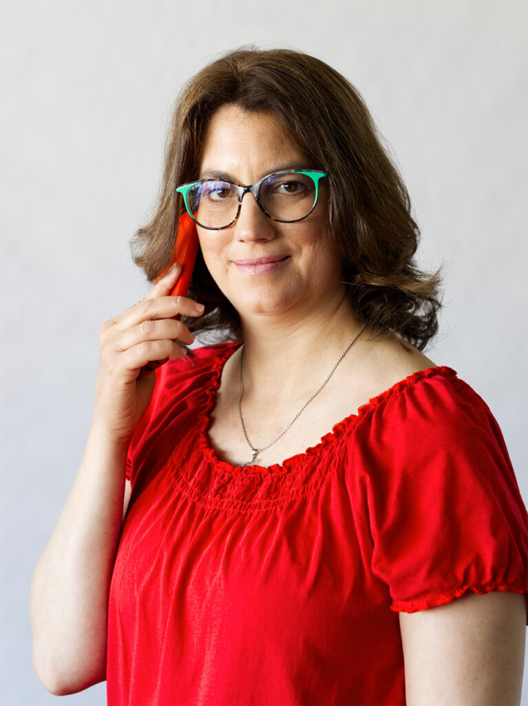Ez egy fotó: Merényi Katalin Gabriella matematika tanár, life és business coach, art coach telefonál. A hölgy ránk mosolyog, piros mobiltelefonját a füléhez tartja. Haja barna, hullámos, vállig ér. Szemüvegkerete zöld-barna. Piros rövidujjú felsőt visel, nyakában egy ezüst kolibri medál, mint a gondolat felszabadulásának szimbóluma.